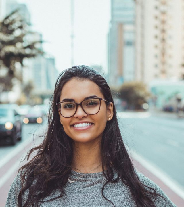 A young woman with glasses smiling on a city street, embracing urban lifestyle.